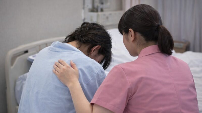 A midwife gently supporting a mother in a hospital room, seen from behind
